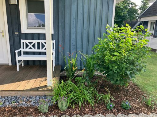 Eingang mit einer weißen Bank und bunten Pflanzen neben einem blauen Holzhaus.