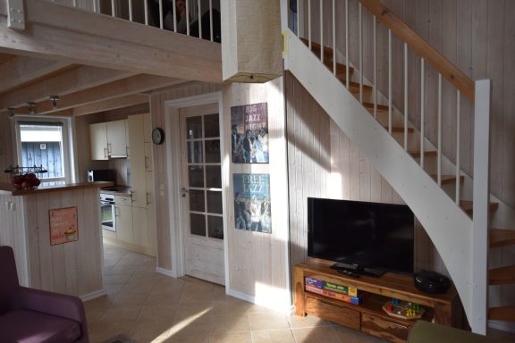 Helles Wohnzimmer mit Treppe, Küchenecke und Fernseher auf Holzständermöbel.